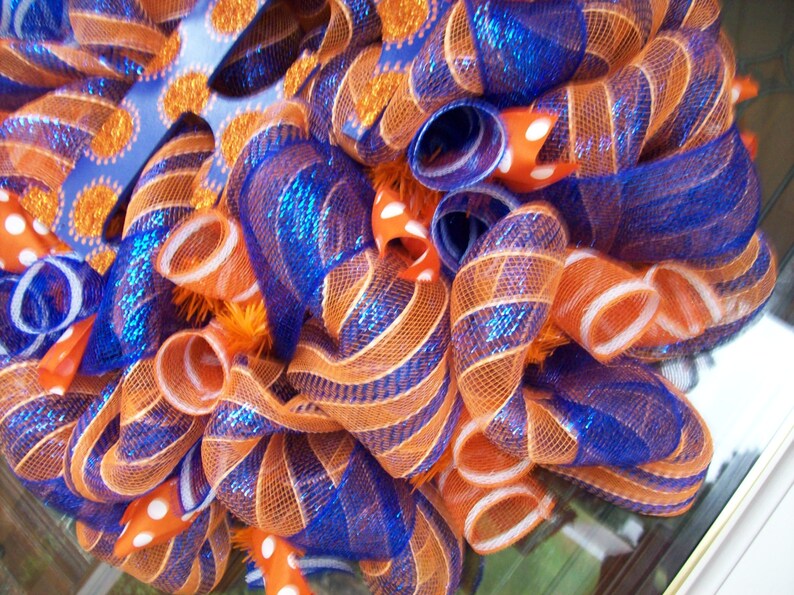 May include: A wreath made of orange and blue mesh ribbon with white polka dots. The wreath is decorated with a blue and orange glitter bow.