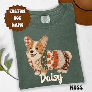 Może przedstawiać: Koszulka w kolorze mchu z ilustracją psa rasy Corgi w stylu patchwork, z różnymi jesiennymi wzorami, w tym dyniami, liśćmi i kratą. Imię Daisy jest nadrukowane na biało pod psem. Brązowa etykieta zawiera napis CUSTOM DOG NAME.