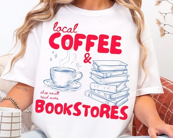 May include: A white short-sleeve t-shirt featuring a graphic print with the red text "local COFFEE & BOOKSTORES" and the smaller red text "shop small read more". The design includes a blue line-art illustration of a coffee cup and a stack of books.