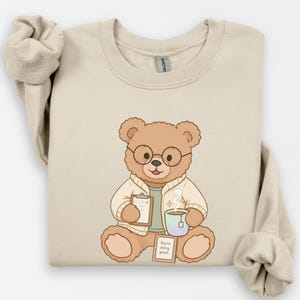 Peut inclure: Sweat-shirt beige avec un ours en peluche de dessin animé portant des lunettes et une blouse de laboratoire. L'ours tient un presse-papiers et une tasse de thé fumante. Un panneau indique "You're doing great."