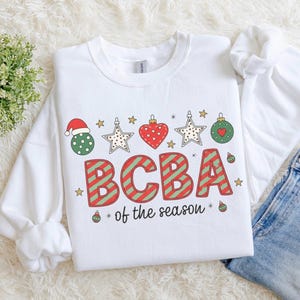 Op de afbeelding: Witte crewneck sweatshirt met een feestelijk ontwerp. Het ontwerp bevat een kerstmuts, sterren, harten en ornamenten boven de tekst "BCBA of the season" in rood en groen. Een takje groen is linksboven zichtbaar.