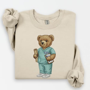 May include: A beige sweatshirt featuring a cartoon bear dressed as a medical professional. The bear wears teal scrubs and holds a rolled document and a clipboard. The text "PT BEAR" is on the scrubs. The bear has a friendly expression.