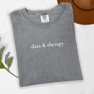 Könnte beinhalten: Graues Comfort Colors T-Shirt mit dem weißen Aufdruck "data & therapy". Das Shirt ist gefaltet und hat einen Rundhalsausschnitt. Ein brauner Hut ist teilweise im Hintergrund zu sehen.