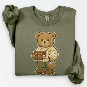 May include: Olive green sweatshirt featuring an embroidered teddy bear design. The bear is wearing a sweater and holding a star-studded board and a basket. The bear is wearing green pants and brown shoes. The sweatshirt has long sleeves.