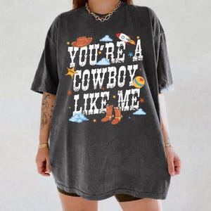 May include: A charcoal gray short-sleeve t-shirt featuring the white distressed text "YOU'RE A COWBOY LIKE ME" surrounded by colorful graphics including a brown cowboy hat, a rocket ship, a yellow ball, and brown cowboy boots with blue clouds.