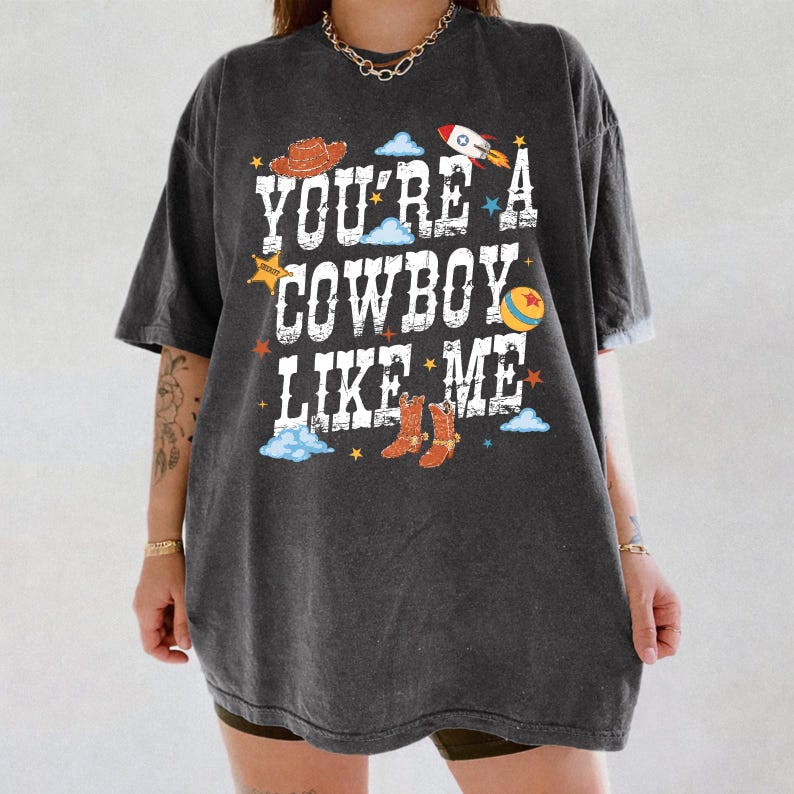 May include: A charcoal gray short-sleeve t-shirt featuring the white distressed text "YOU'RE A COWBOY LIKE ME" surrounded by colorful graphics including a brown cowboy hat, a rocket ship, a yellow ball, and brown cowboy boots with blue clouds.