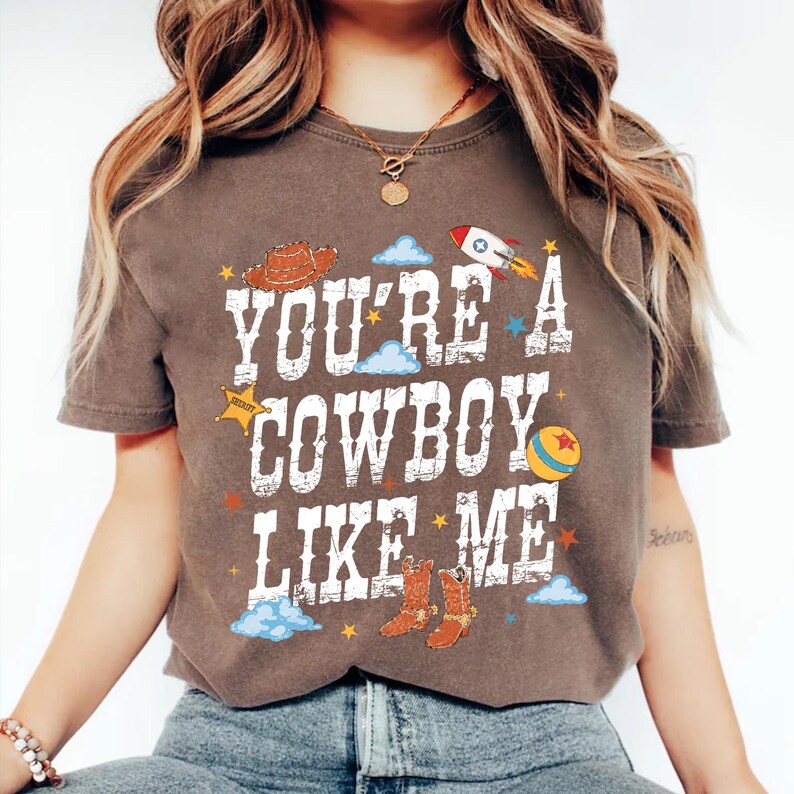 May include: A dark brown short-sleeve t-shirt featuring the white distressed text "YOU'RE A COWBOY LIKE ME" surrounded by colorful graphic illustrations of a cowboy hat, a rocket ship, a sheriff star, a ball, and cowboy boots with blue clouds.