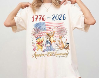 May include: A cream-coloured t-shirt with a patriotic design. It features the text "1776-2026" in red and blue, an American flag, fireworks, and cartoon characters. The text "America - 250th Anniversary" is written in gold.