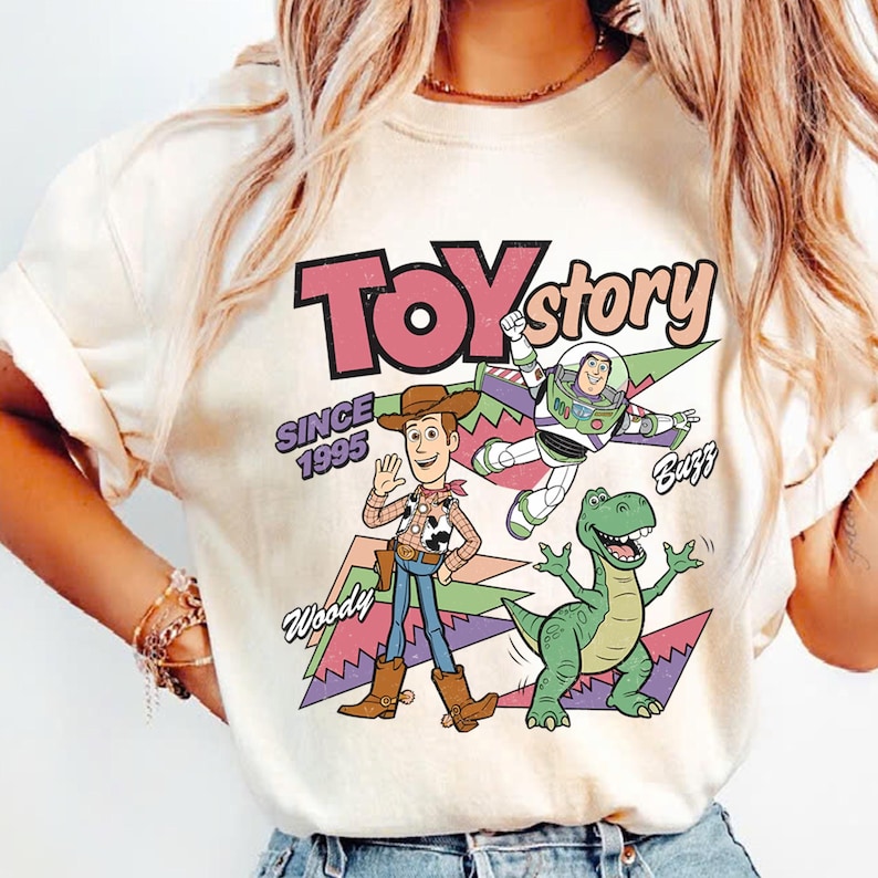 May include: Cream-coloured t-shirt with a vibrant graphic of Toy Story characters Woody, Buzz Lightyear, and Rex. The text "Toy Story" is in pink and black, with "Since 1995" below. The shirt has short sleeves.