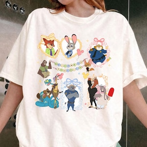 May include: White t-shirt with a colourful cartoon design featuring characters from Zootopia, such as a fox and a bunny, within decorative frames. The text "WELCOME TO ZOOTOPIA" is displayed across the top. A playful, whimsical design.