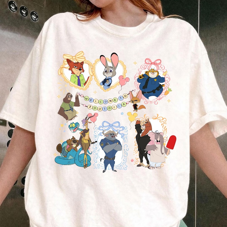 May include: White t-shirt with a colourful cartoon design featuring characters from Zootopia, such as a fox and a bunny, within decorative frames. The text "WELCOME TO ZOOTOPIA" is displayed across the top. A playful, whimsical design.