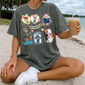 May include: Dark grey t-shirt featuring framed cartoon characters. The shirt showcases characters like a fox, rabbit, and others, set against a beach backdrop. The design includes decorative frames and a string of colourful circles.