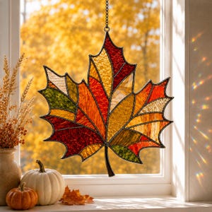 May include: A stained glass maple leaf suncatcher featuring a mosaic of textured glass panels in shades of red, orange, yellow, and green, outlined in dark metal solder, suspended by a thin chain against a bright window with autumn foliage.