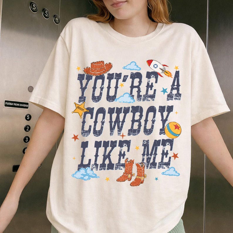 May include: A cream-colored short-sleeve t-shirt featuring the dark blue distressed text "YOU'RE A COWBOY LIKE ME" surrounded by colorful graphics including a brown cowboy hat, a rocket ship, a yellow sheriff star, a ball, and orange cowboy boots.