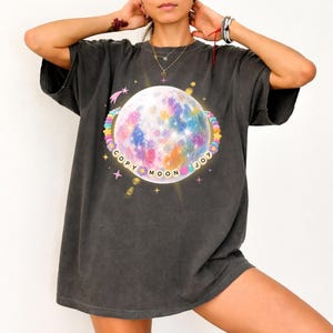 May include: Dark gray t-shirt featuring a colorful moon graphic with the words "COPY MOON JOY" in a circular design. The moon is surrounded by a ring of pastel-colored beads and stars. The shirt has short sleeves and a relaxed fit.