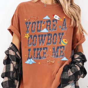 May include: A rust-colored short-sleeve t-shirt featuring the text "YOU'RE A COWBOY LIKE ME" in distressed blue lettering. The graphic includes illustrations of a brown cowboy hat, a rocket ship, a yellow ball, and cowboy boots with stars.
