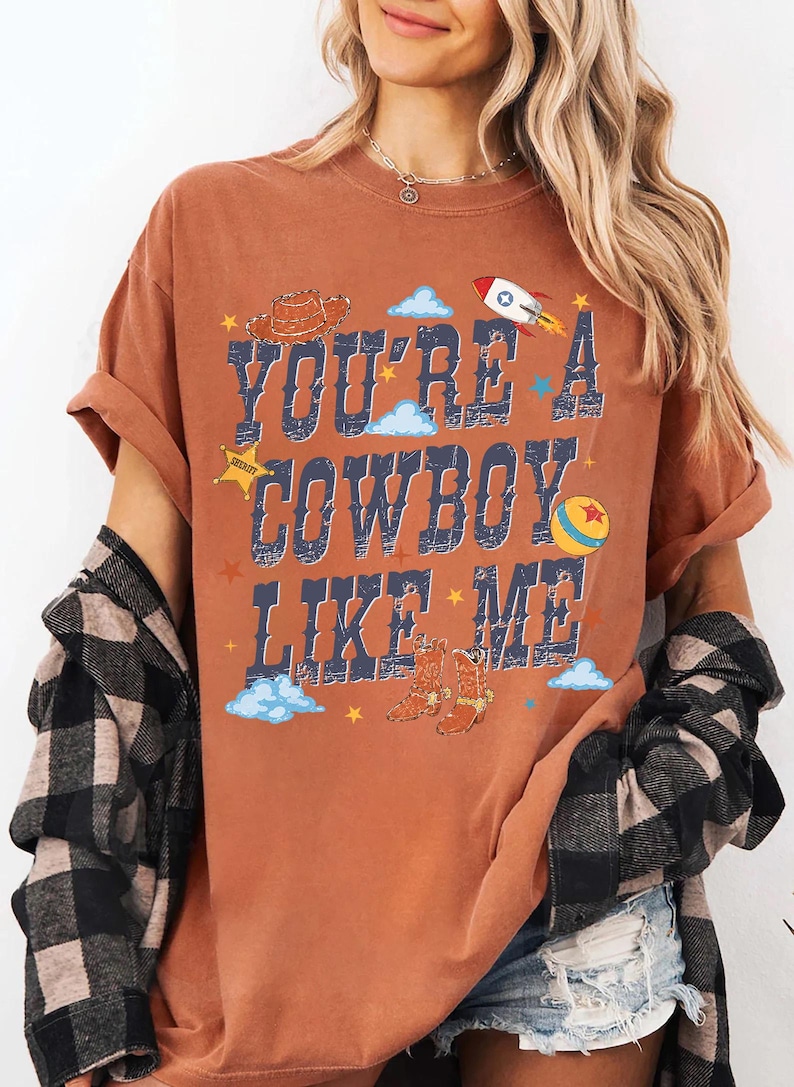 May include: A rust-colored short-sleeve t-shirt featuring the text "YOU'RE A COWBOY LIKE ME" in distressed blue lettering. The graphic includes illustrations of a brown cowboy hat, a rocket ship, a yellow ball, and cowboy boots with stars.