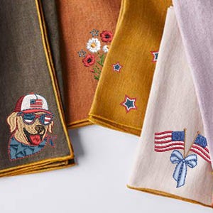 Puede incluir: Primer plano de varias toallas decorativas dobladas. Una toalla presenta un perro bordado con gafas de sol y un sombrero con la bandera estadounidense. Otras toallas tienen flores, estrellas y banderas americanas bordadas.