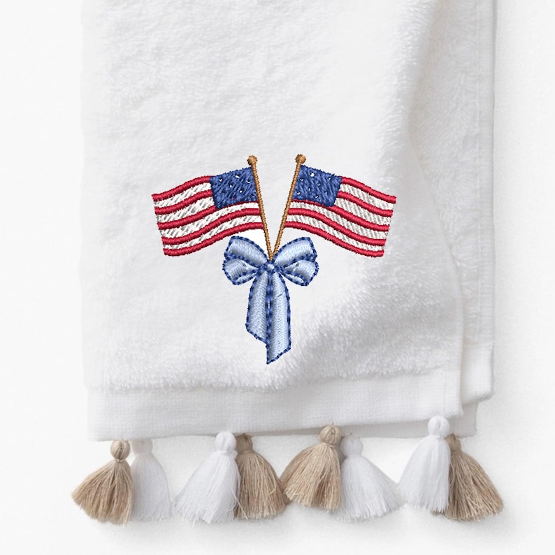 Puede incluir: Toalla blanca con banderas americanas bordadas y un lazo azul. Las banderas tienen rayas rojas y blancas y campos de estrellas azules. La toalla est&aacute; rematada con borlas blancas y beige alternas.