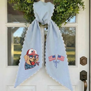 Puede incluir: Bufanda decorativa azul claro con bordes festoneados blancos. Presenta bordados de un perro con gafas de sol y gorra de b&eacute;isbol, y dos banderas americanas. La bufanda est&aacute; atada a una corona verde.