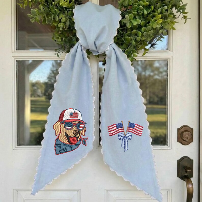 Puede incluir: Bufanda decorativa azul claro con bordes festoneados blancos. Presenta bordados de un perro con gafas de sol y gorra de b&eacute;isbol, y dos banderas americanas. La bufanda est&aacute; atada a una corona verde.