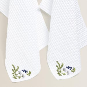 May include: Two white waffle-weave towels with embroidered floral designs. The towels feature a corner design with green leaves, blue berries, and white flowers. The towels are hanging against a neutral background.