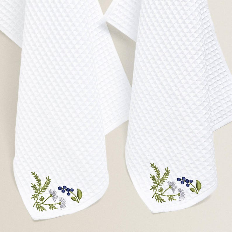 May include: Two white waffle-weave towels with embroidered floral designs. The towels feature a corner design with green leaves, blue berries, and white flowers. The towels are hanging against a neutral background.