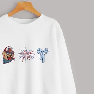 Puede incluir: Sudadera blanca con dise&ntilde;os patri&oacute;ticos bordados. Presenta un golden retriever con gafas de sol y un sombrero con un dise&ntilde;o de bandera estadounidense, una explosi&oacute;n de fuegos artificiales y un lazo azul.