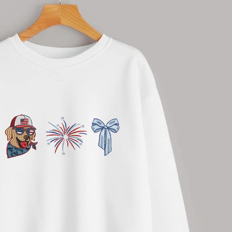 Puede incluir: Sudadera blanca con dise&ntilde;os patri&oacute;ticos bordados. Presenta un golden retriever con gafas de sol y un sombrero con un dise&ntilde;o de bandera estadounidense, una explosi&oacute;n de fuegos artificiales y un lazo azul.