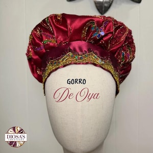 May include: A deep red satin headpiece featuring intricate gold embroidery, multicolored sequins, and beaded accents. The fabric is gathered at the top, and the front band displays the text GORRO De Oya. A circular logo with the text DIOSA'S CREATIONS is visible.