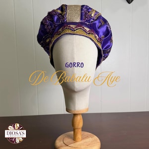 Peut inclure: Un couvre-chef en satin violet orné de garnitures à paillettes dorées et d'un panneau central texturé doré. L'accessoire est présenté sur une tête de mannequin. Le texte GORRO De Babalu Aye et le logo DIOSA'S CREATIONS sont visibles.
