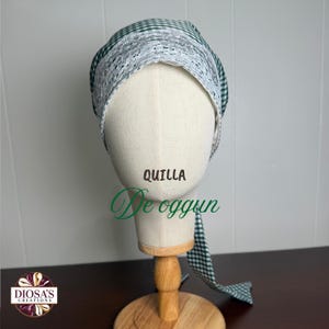 May include: A green and white gingham head covering featuring a wide band of white floral lace trim along the front edge. The fabric has a tie-back design with long tails. The text "QUILLA" and "De oggun" are printed on the front of the head covering.