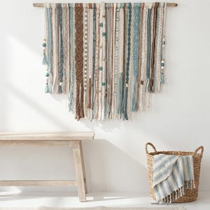 May include: A woven wall hanging with a wooden rod, featuring vertical strands in shades of blue, brown, and cream. Some strands have decorative beads and tassels. A light wooden bench and a woven basket with a blue and white striped throw are in the foreground.