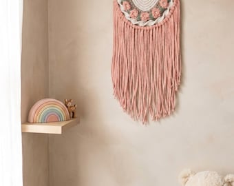 May include: A pink macrame wall hanging with a floral design and fringe, displayed on a light-colored wall. A wooden rainbow and deer figurine sit on a shelf. A large teddy bear is in the corner.