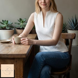 May include: A sleeveless white top with a vertical ribbed texture and a V-neck. The person is seated at a wooden table, holding a mug. The background includes potted plants and a wooden chair. The logo for "The Closette" is in the lower left corner.