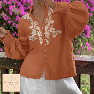 Puede incluir: Una blusa naranja con bordados florales blancos y mangas largas y abullonadas. La parte superior con cuello en V presenta pequeños botones dorados en la parte delantera. La blusa se combina con pantalones blancos. Se ve el logo "The Closette Premium".