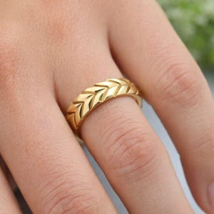 May include: A gold-colored ring with a textured chevron pattern. The ring is worn on a finger and appears to be made of metal. The design features a series of repeating, pointed shapes that create a textured surface.