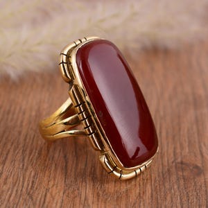 Può includere: Un anello color oro con una grande pietra preziosa rettangolare lucida, rosso scuro. L'anello ha un'incastonatura dettagliata e strutturata attorno alla pietra e sulla fascia. L'anello è esposto su una superficie di legno.