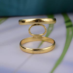May include: A gold-colored ring with a unique design. The ring features a band, an oval-shaped loop, and a horizontal bar. The ring is made of a shiny, metallic material. The ring's design is modern and geometric.