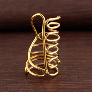 May include: A gold-colored metal hair clip with a unique spiral design. The clip features a series of curved lines and a coiled section, creating an open, airy structure. The clip is designed to hold hair in place.