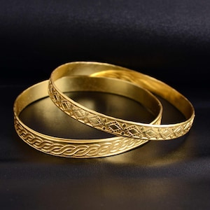 May include: Two gold bangles with intricate patterns. One bangle features a repeating diamond design, while the other has a braided rope-like pattern. The bangles are displayed on a dark surface, highlighting their golden color.