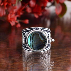 Peut inclure: Une large bague en argent avec un centre en coquille d'ormeau. La bague a une bande détaillée et ornée de motifs et un sertissage articulé pour l'ormeau. La coquille d'ormeau affiche des teintes vertes et bleues irisées. La bague est présentée sur une surface réfléchissante.