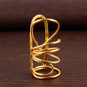 Puede incluir: Un brazalete de metal dorado abstracto. El brazalete presenta una serie de diseños curvos en forma de espiral. El brazalete tiene una superficie texturizada y se muestra sobre un fondo marrón oscuro.