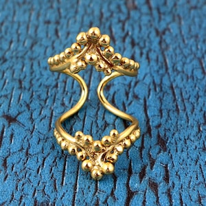 Könnte beinhalten: Ein goldfarbener Ring mit einem einzigartigen Design. Der Ring hat zwei verbundene Bänder, die jeweils mit kleinen, kugelförmigen Verzierungen versehen sind. Die oberen und unteren Abschnitte haben eine blütenartige Anordnung der Kugeln. Der Ring ist vor einem blauen Hintergrund platziert.