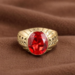 Op de afbeelding: Een goudkleurige ring met een ovale, gefacetteerde, rode edelsteen. De ring heeft een decoratieve band met uitgesneden details. De rode edelsteen is het middelpunt, tegen een bruine achtergrond.