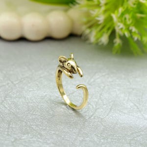May include: A gold-colored ring featuring a mouse design. The ring has an open design, with the mouse's head and paws forming the top and the band curving around to meet the tail. The ring is made of a shiny, metallic material.