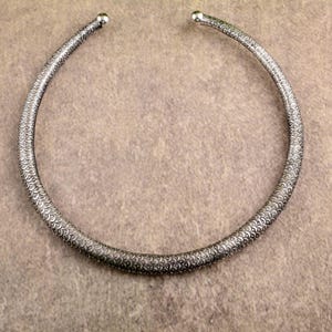 Puede incluir: Un collar gargantilla plateado con un patrón texturizado en forma de escamas. El diseño abierto presenta pequeños terminales redondeados en cada extremo. El collar se muestra sobre una superficie neutra de color beige.