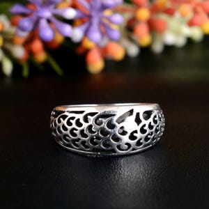 Puede incluir: Un anillo plateado con un diseño calado. El anillo presenta una banda ancha con un patrón repetitivo de recortes en espiral. El anillo está sobre un fondo oscuro con flores coloridas en el fondo.