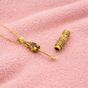 Puede incluir: Un collar de tono dorado con una cadena fina y un colgante que presenta un componente de varilla delgada y extendida. Junto al colgante se encuentra una tapa cilíndrica dorada separada con un patrón trenzado texturizado.