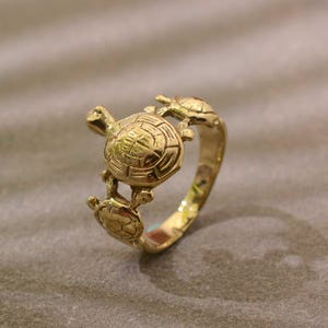 Può includere: Un anello color oro con tre disegni di tartarughe. L'anello presenta un design dettagliato di tartaruga al centro, con due tartarughe più piccole ai lati. L'anello è realizzato in un materiale metallico lucido.